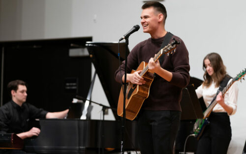 Three students on stage playing a piano, guitar and bass.