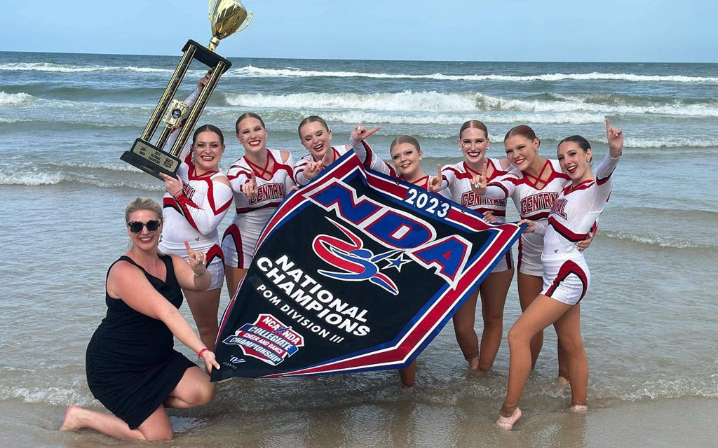 Central dance team on beach holding championship banner and trophy